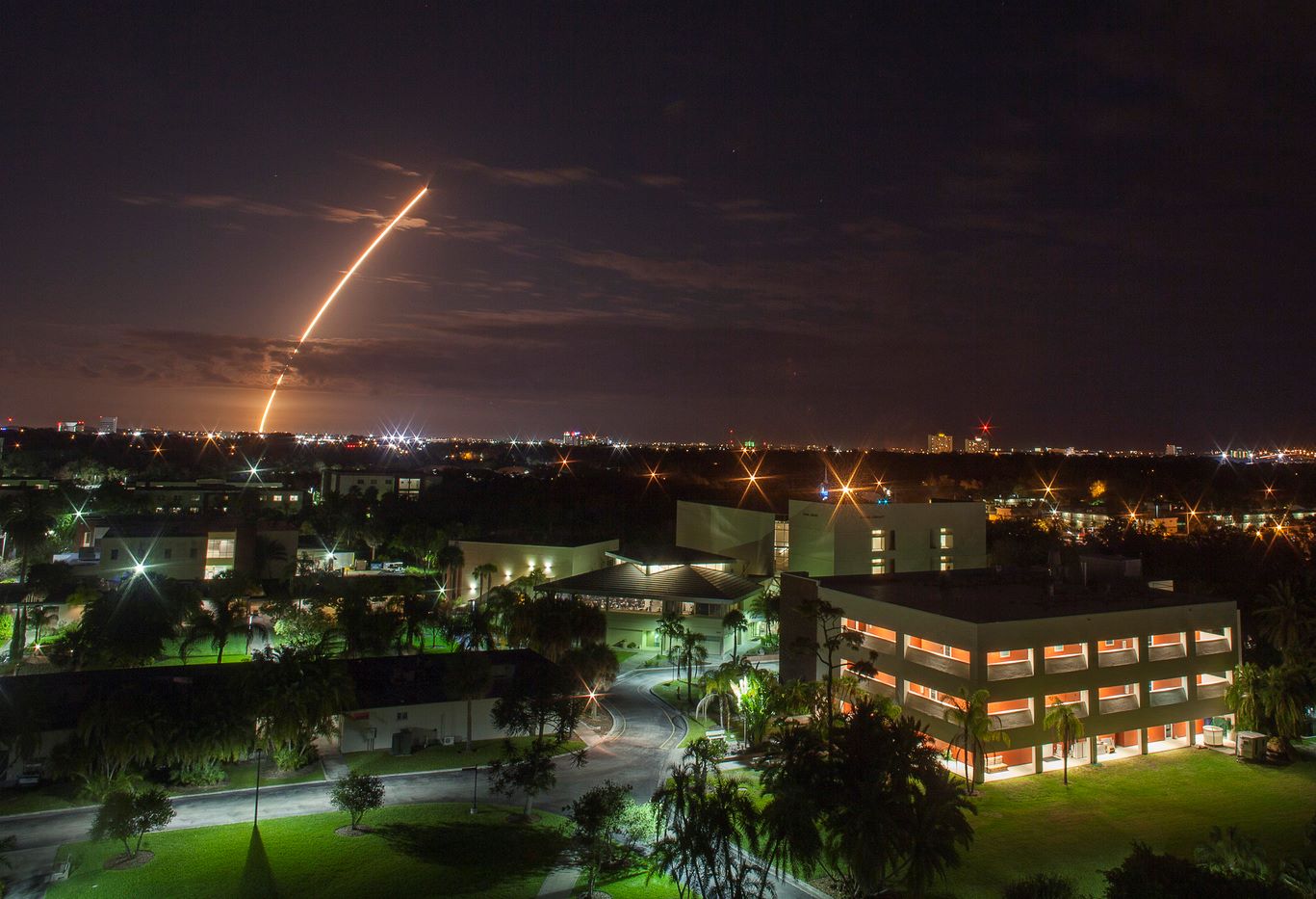 Nighttime view of a rocket launch seen over a university campus, with the trajectory illuminated against a dark sky and campus buildings lit up below